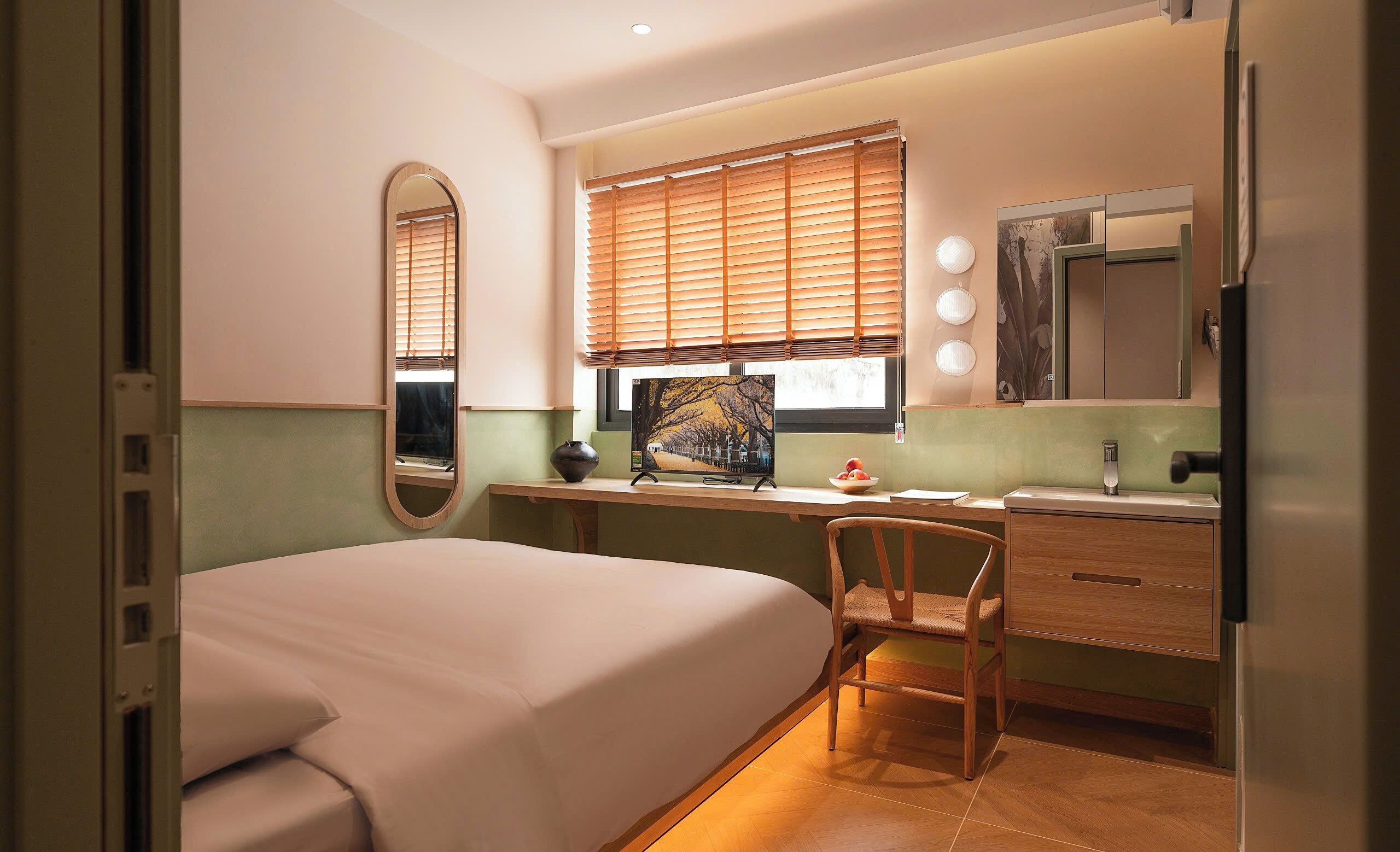A bedroom with a bed, a desk, a chair, a mirror, and a window with blinds.