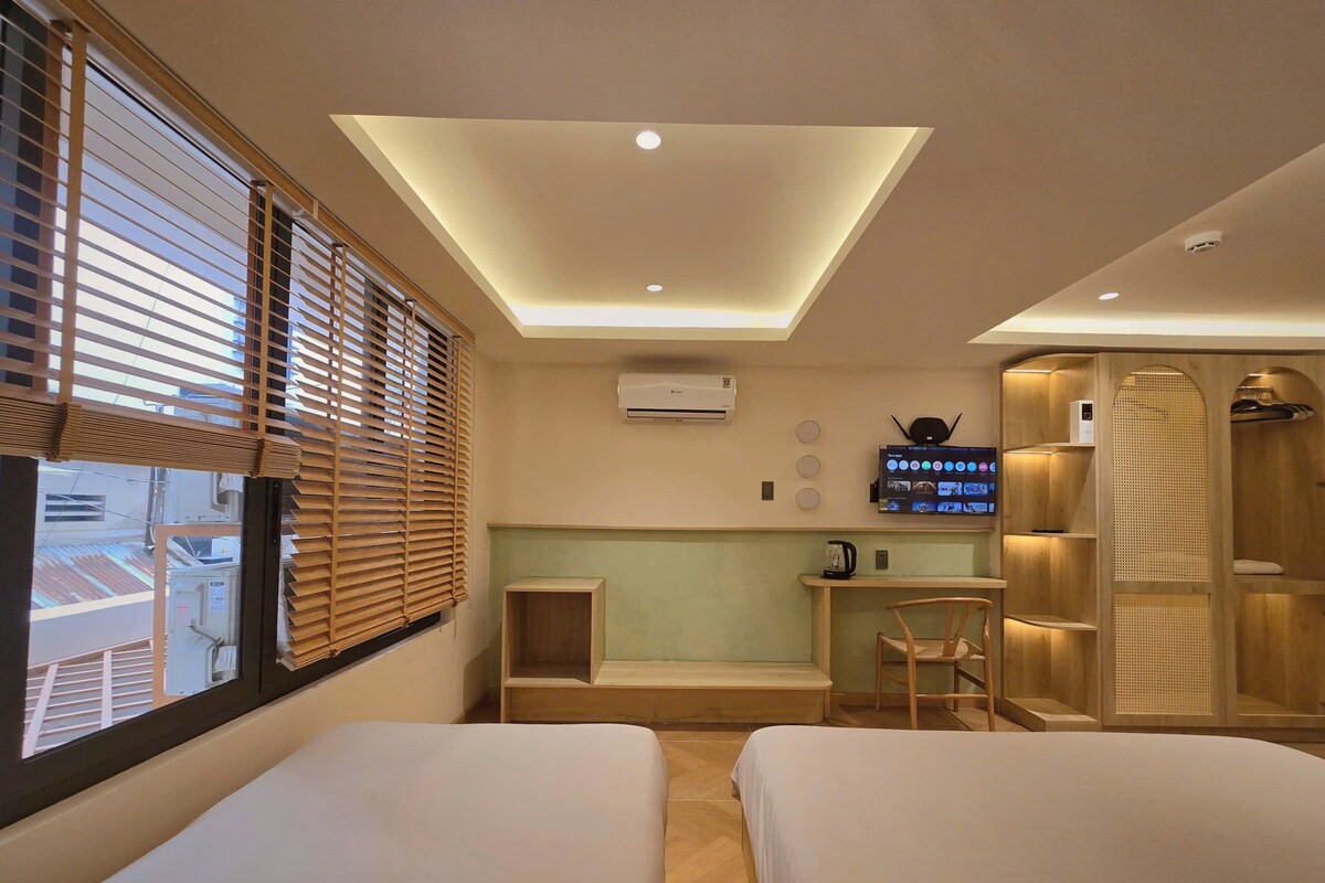 A hotel room with two beds, a desk, a TV, and a shower area.