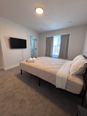 Une chambre à coucher comprenant un lit, un téléviseur à écran plat fixé au mur, une fenêtre avec des rideaux et une porte donnant sur une autre pièce.