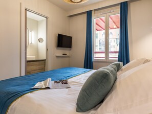 Et hotelværelse med seng, fjernsyn, et vindue med gardiner og udsigt til bygninger.