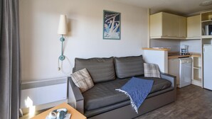 Ein kompakter Wohnbereich mit einer Couch, einem Tisch mit Lampe, einer Küchenzeile und einem Poster an der Wand.