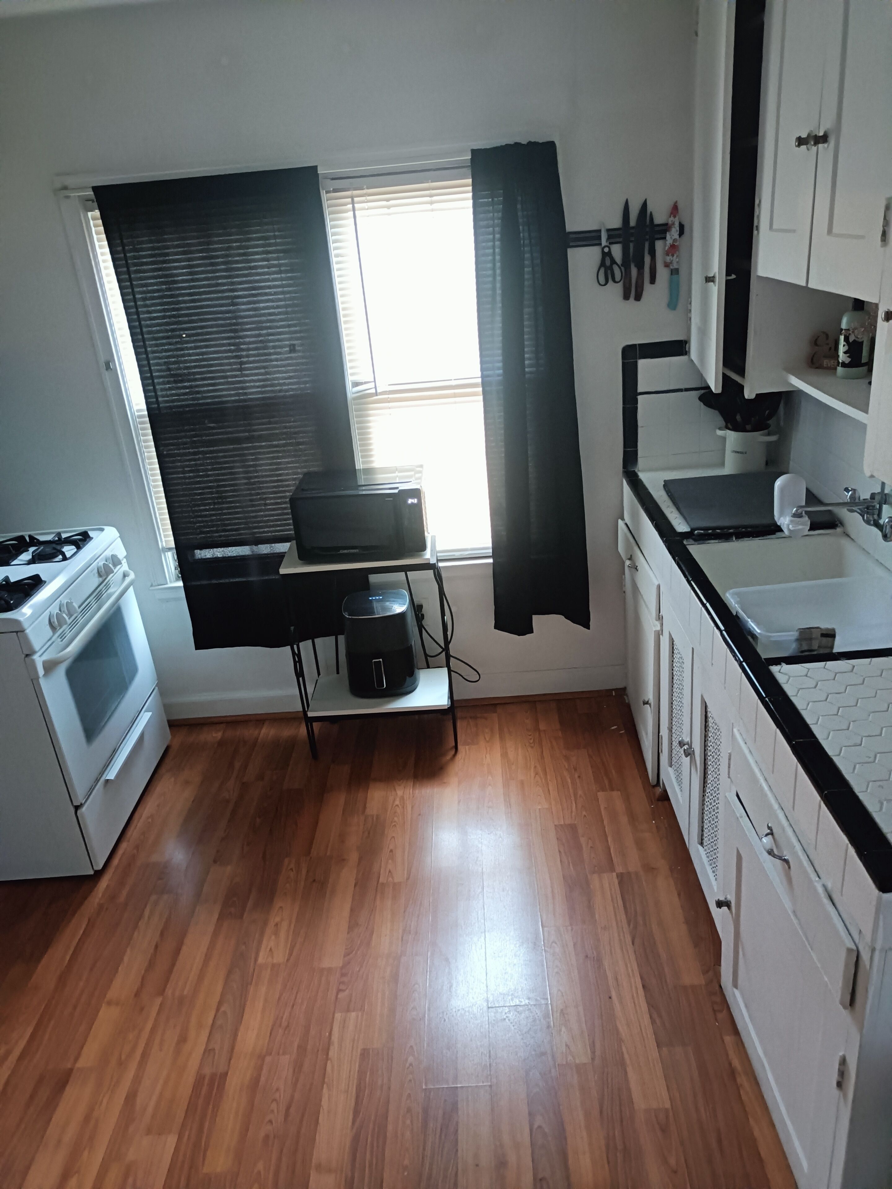 A kitchen with a stove, oven, and a coffee maker on a cart, with a window and curtains.
