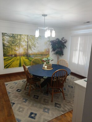 A dining area with a round table, four chairs, a chandelier, a large painting, and a potted plant.