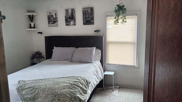Un dormitorio con cama, mesita de noche, ventana con persianas, estante en la pared con una planta y pared con tres cuadros enmarcados.