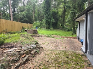 Ein gepflasterter Patio-Bereich mit einem Ziegelweg, der zu einem hölzernen Geräteschuppen führt, umgeben von einem Holzzaun und üppiger Vegetation.