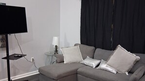 Una sala de estar moderna con un sofá seccional gris, un televisor de pantalla plana sobre un mueble bajo, una mesita auxiliar con una lámpara y una cortina negra.