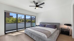 Ein modernes Schlafzimmer mit einem großen Bett, einem Deckenventilator, einem Nachttisch und Blick auf Bäume und Berge durch große Fenster.