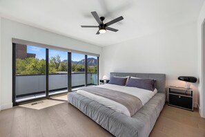 A modern bedroom with a large bed, a ceiling fan, a nightstand, and a view of trees and mountains through large windows.
