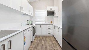 A compact kitchen with white cabinetry, a stainless steel refrigerator, a microwave, and a window with blinds. - Dalton Bungalow - Heritage Character (Orange)