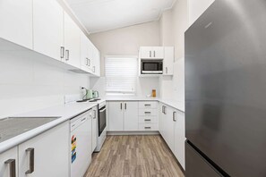 A compact kitchen with white cabinetry, a stainless steel refrigerator, a microwave, and a window with blinds. - Dalton Bungalow - Heritage Character (Orange)