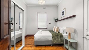 A bedroom with a bed, a bedside table with a lamp, a window with blinds, and a mirror. - Dalton Bungalow - Heritage Character (Orange)
