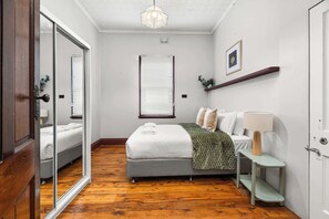 A bedroom with a bed, a bedside table with a lamp, a window with blinds, and a mirror. - Dalton Bungalow - Heritage Character (Orange)