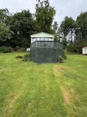 Un pequeño espacio al aire libre delimitado por una cerca, con una estructura de techo verde, rodeado de césped y árboles.