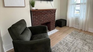 Ein Wohnzimmer mit Kamin aus Backsteinen, einem flachbildfernseher darüber montiert, einem dunklen Ohrensessel und einem gemusterten Teppich.