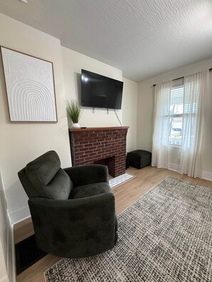 Ein Wohnzimmer mit Kamin aus Backsteinen, einem flachbildfernseher darüber montiert, einem dunklen Ohrensessel und einem gemusterten Teppich.