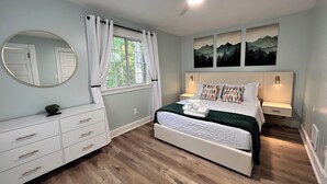 Ein Schlafzimmer mit einem Bett, einer Kommode, einem Spiegel, einem Fenster mit Vorhängen, einem Deckenventilator und Wandleuchten.