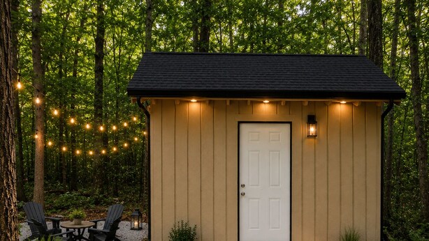 Une petite cabane en bois avec une porte blanche, entourée d’un chemin de gravier, de plantes en pot et de guirlandes lumineuses.