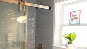 Et badeværelse med brusekabine i glas, toilet, vaskebord med skuffer samt et vindue med en plante.