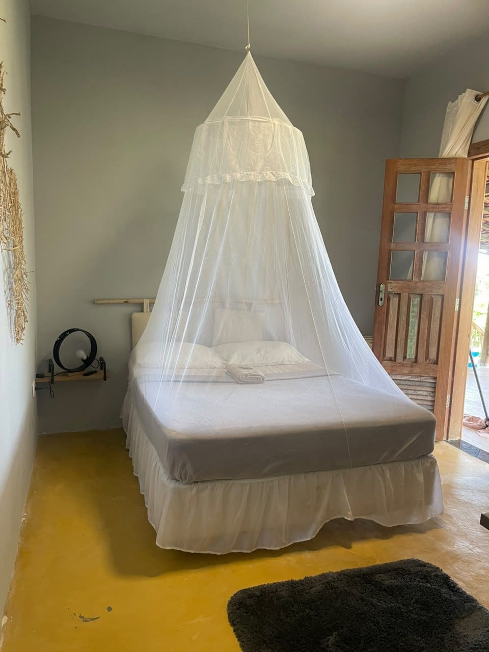 Un llit individual amb un dosser blanc a una habitació amb una porta de fusta i un sòl groc.