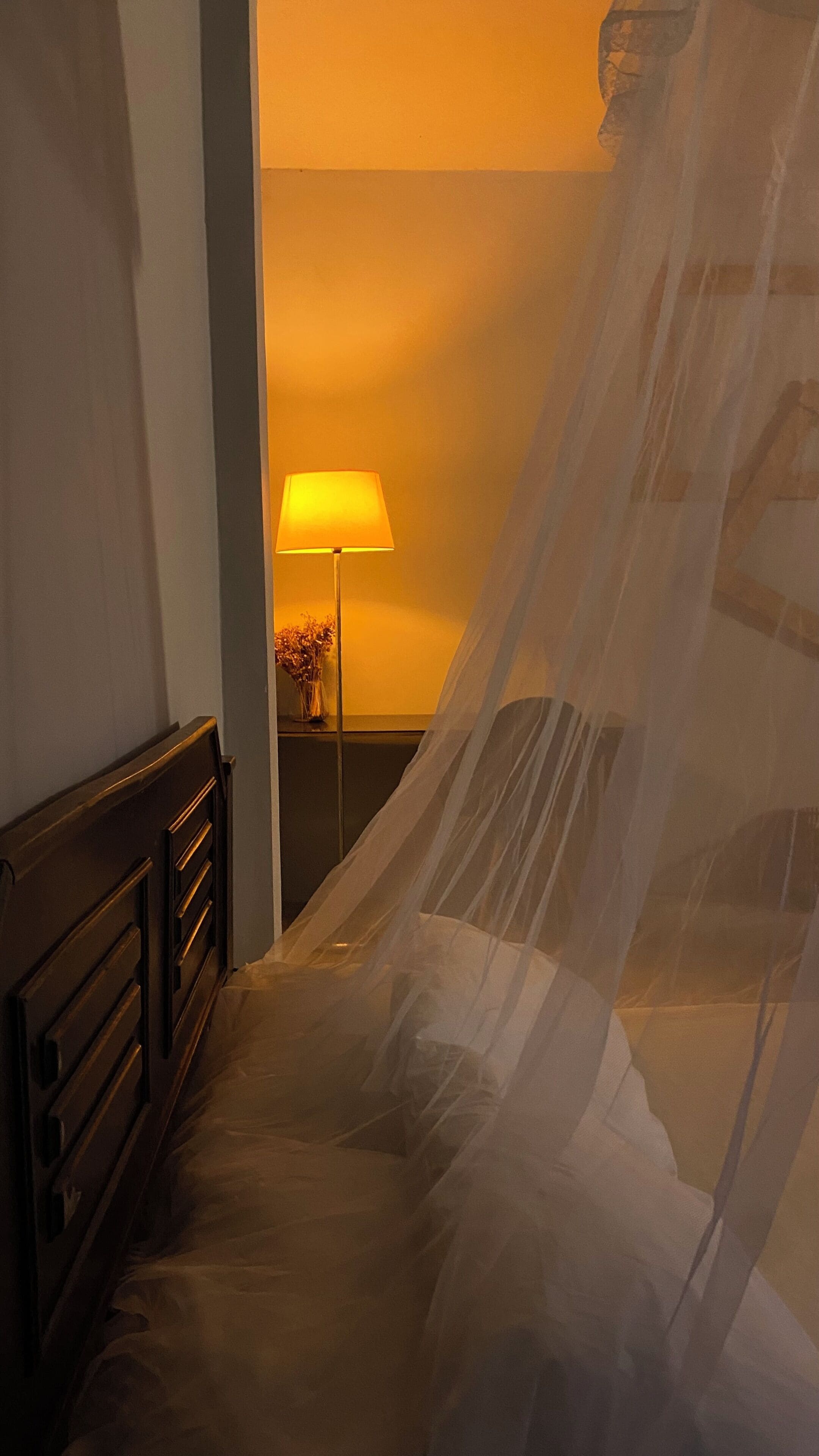 Quarto com cama, mesa de cabeceira, abajur e cortina translúcida.