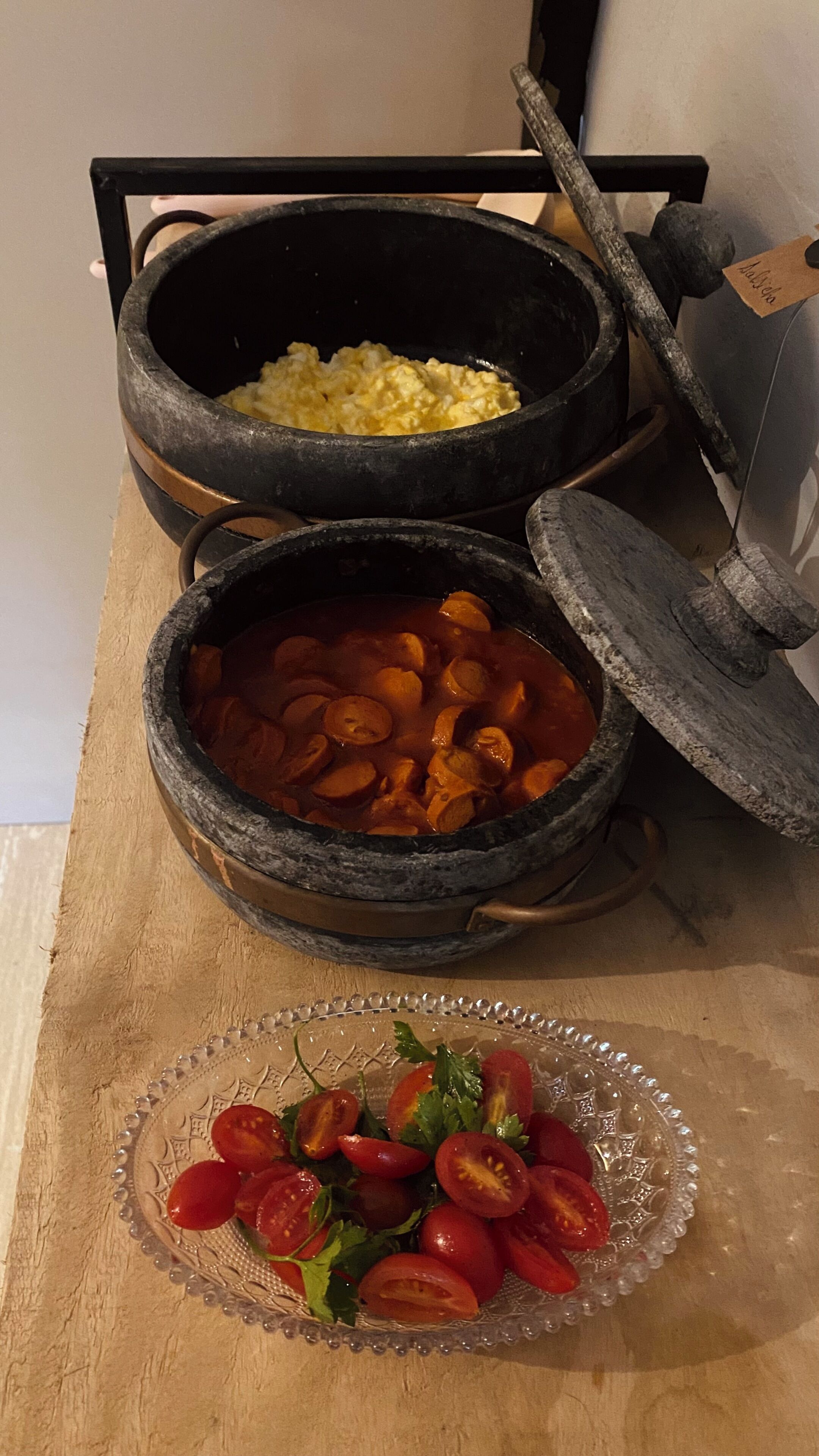Dos ollas de piedra grandes sobre una estufa, una con tapa, y un plato de vidrio con rodajas de tomate y verduras de hoja verde.