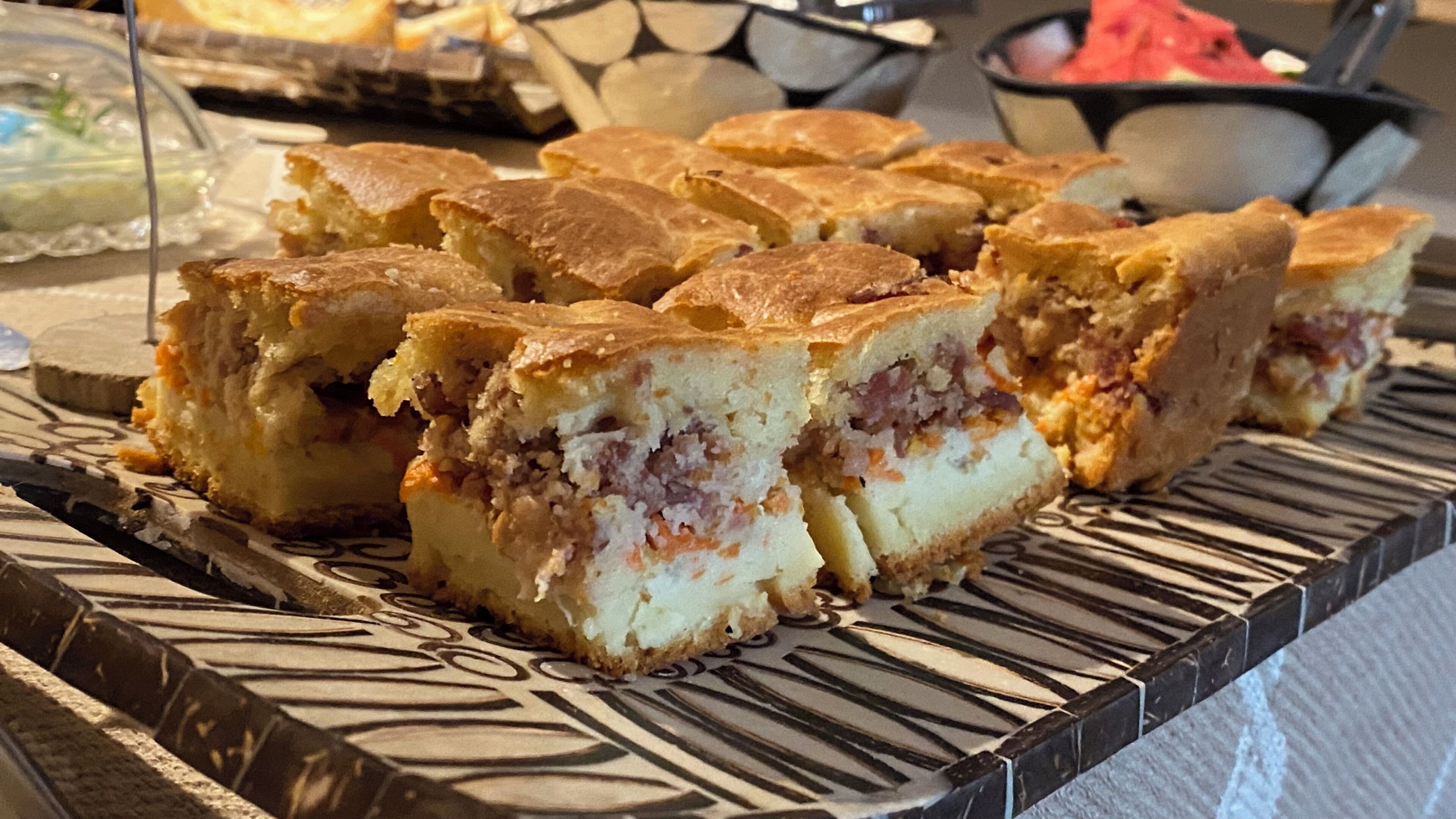 A tray of square pastries with a golden-brown crust, arranged on a patterned serving board.