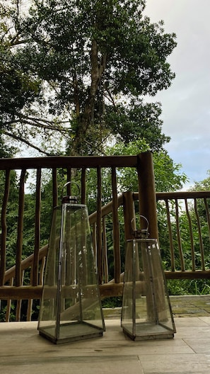 Two clear glass lanterns on a wooden deck with a metal railing and a large tree in the background.