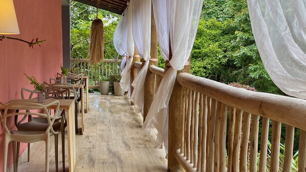 Un área de estar al aire libre con techo, con sillas y mesas de madera, baranda de madera y plantas en macetas.
