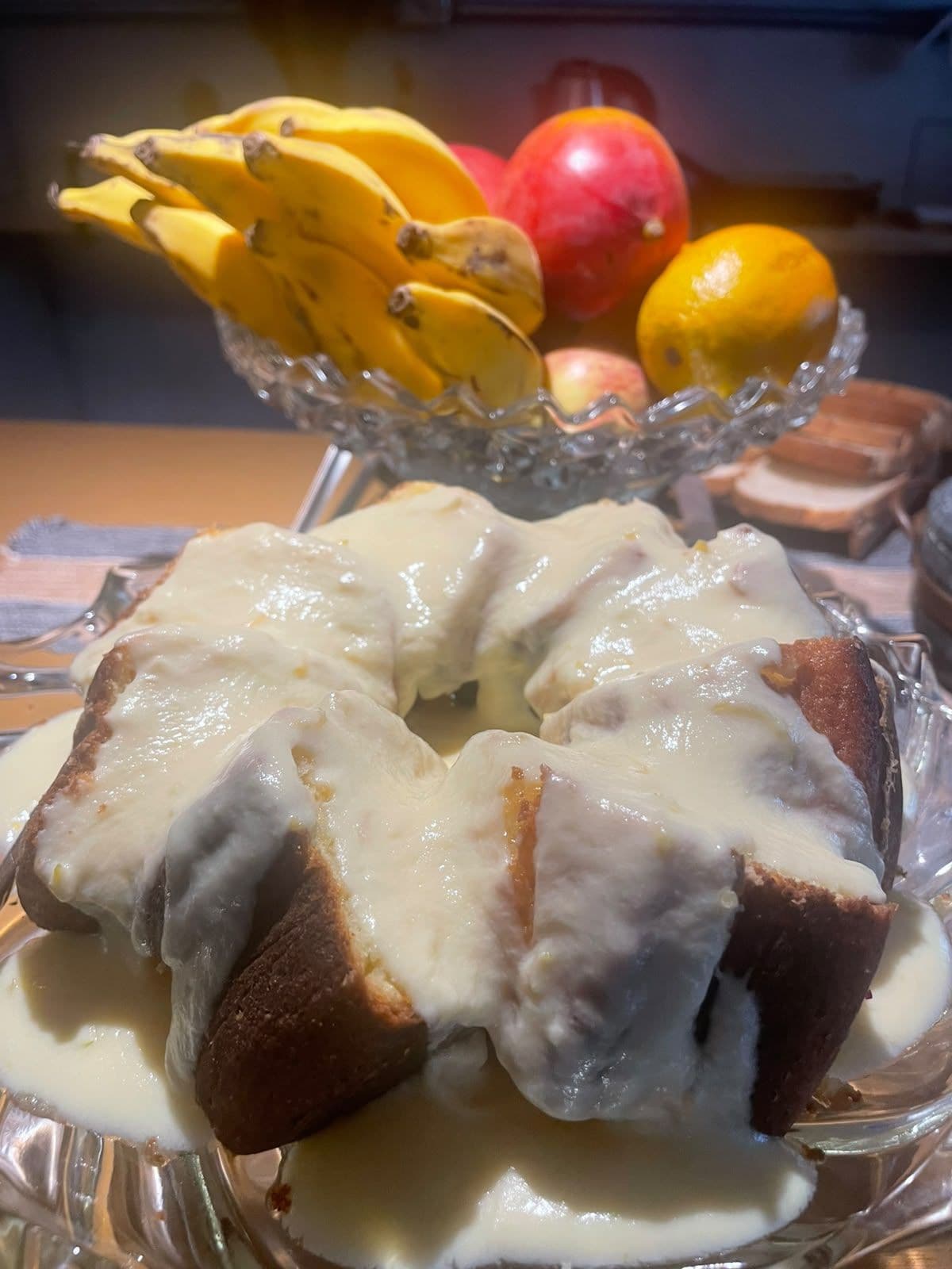 A bundt cake with white icing on a plate, with a bowl of fruit in the background.
