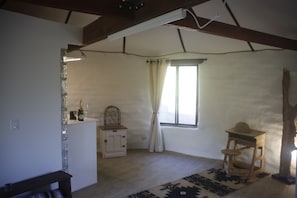 Une pièce avec un plafond en bois, une fenêtre avec des rideaux, une petite table sur laquelle se trouve un chapeau, et un buffet blanc contenant des verres à vin.