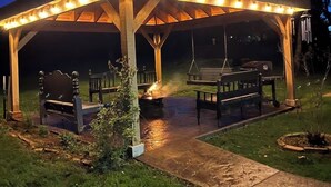 Un espacio al aire libre con techo, bancos y una parrilla, rodeado de césped y árboles.
