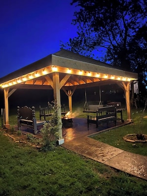 A covered outdoor seating area with benches and a grill, surrounded by grass and trees.