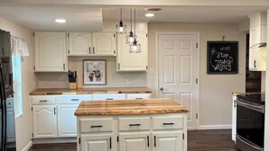 Una cocina amplia con armarios blancos, una isla central, iluminación colgante y una pizarra con una frase motivacional.