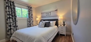 A bedroom with a bed, two bedside tables, a nightstand, a window with curtains, and a wall-mounted artwork.