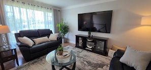 Un salón con un sofá negro, una mesa de centro de vidrio, un televisor en la pared y una planta sobre la mesa.