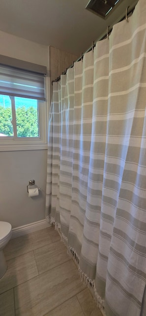 Un baño con cortina de ducha a rayas, inodoro, ventana con vistas a la vegetación y un perchero para toallas.