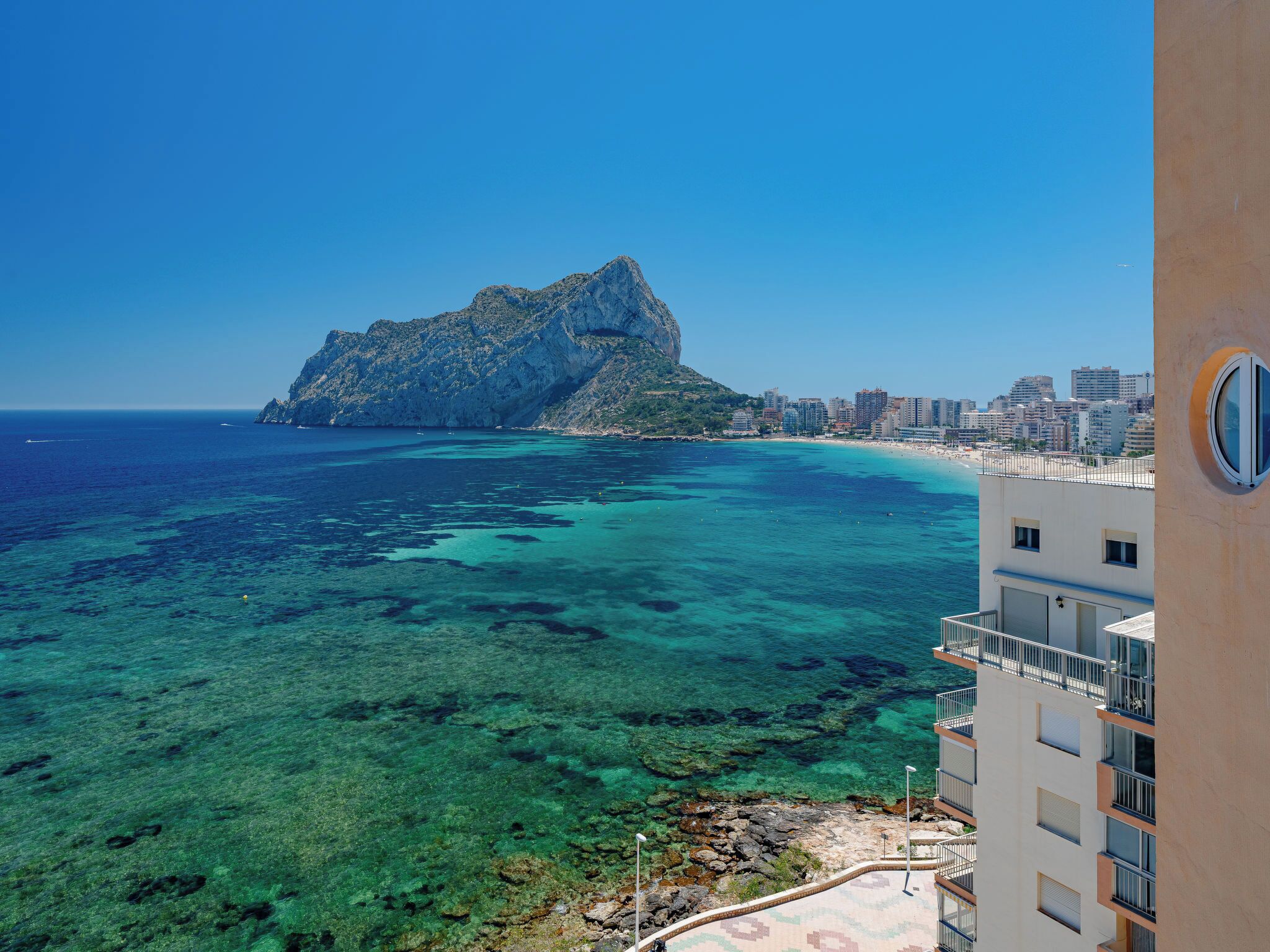 A coastal cityscape with a prominent rocky outcrop, clear blue waters, and a beachfront.