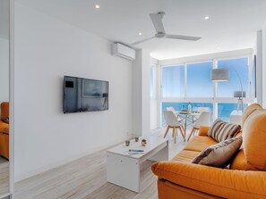 A modern living room with a white sofa, a TV mounted on the wall, a ceiling fan, and a large window offering a view of the sea. - Horizonte by Interhome (Calpe/Calp)