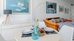 Een moderne woonkamer met een eettafel, een bank en een bed. Er hangen kunstwerken in lijsten aan de muren en er is een plafondventilator.