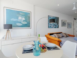 Ein modernes Wohnzimmer mit Esstisch, Sofa und Bett. An den Wänden hängen gerahmte Kunstwerke und eine Deckenleuchte.