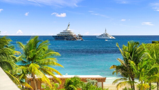 A coastal scene with two large yachts on the water, palm trees, and a building with a balcony. - The Luxe A5 | Beachside @ The Drake Residences (Simpson Bay)