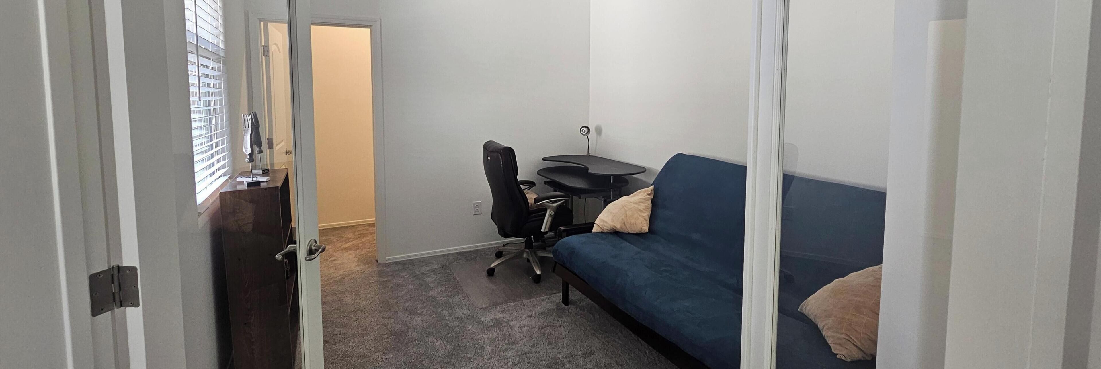 Une pièce avec un canapé bleu, une petite table ronde, un fauteuil de bureau noir et une porte qui donne sur une autre pièce.