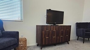 Une pièce avec deux fauteuils sombres, un meuble en bois doté de tiroirs, un téléviseur posé sur le dessus, et une fenêtre avec des stores.