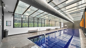 An indoor swimming pool with a glass ceiling, benches along the walls, and a clear view of greenery outside. - Exclusive Stays - SB Grand (Southbank)