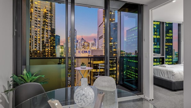 A modern hotel room with a large window offering a city view, a glass table with a vase, a chair, and a bed with a white mattress. - Exclusive Stays - SB Grand (Southbank)