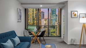 A modern living room with a blue sofa, a dining table with chairs, and a city view through large windows. - Exclusive Stays - SB Grand (Southbank)