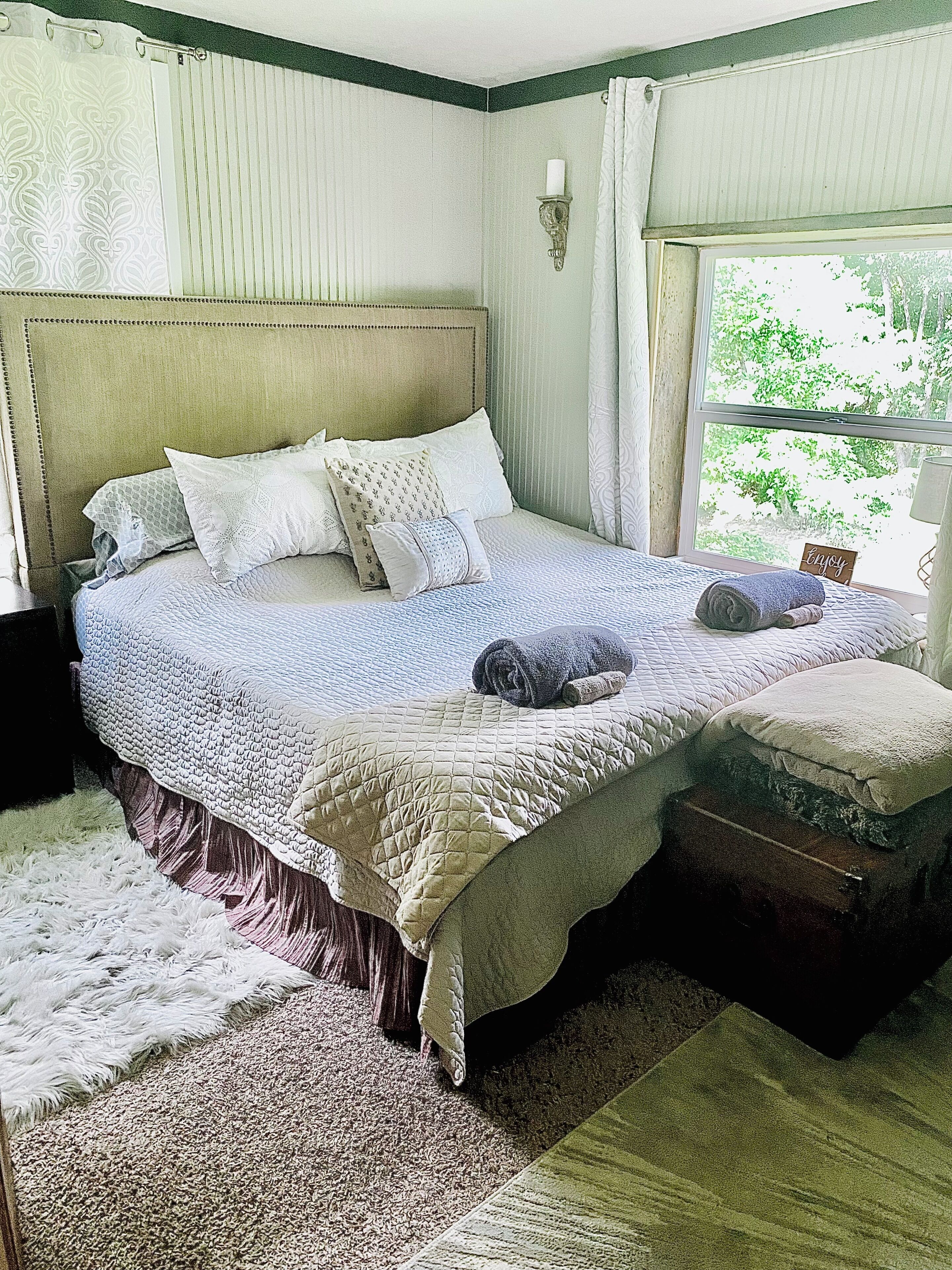 Une chambre avec un grand lit, une table de chevet, une fenêtre avec des rideaux et une vue sur la verdure.