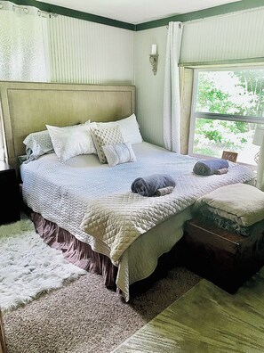 A bedroom with a large bed, a nightstand, a window with curtains, and a view of greenery outside.