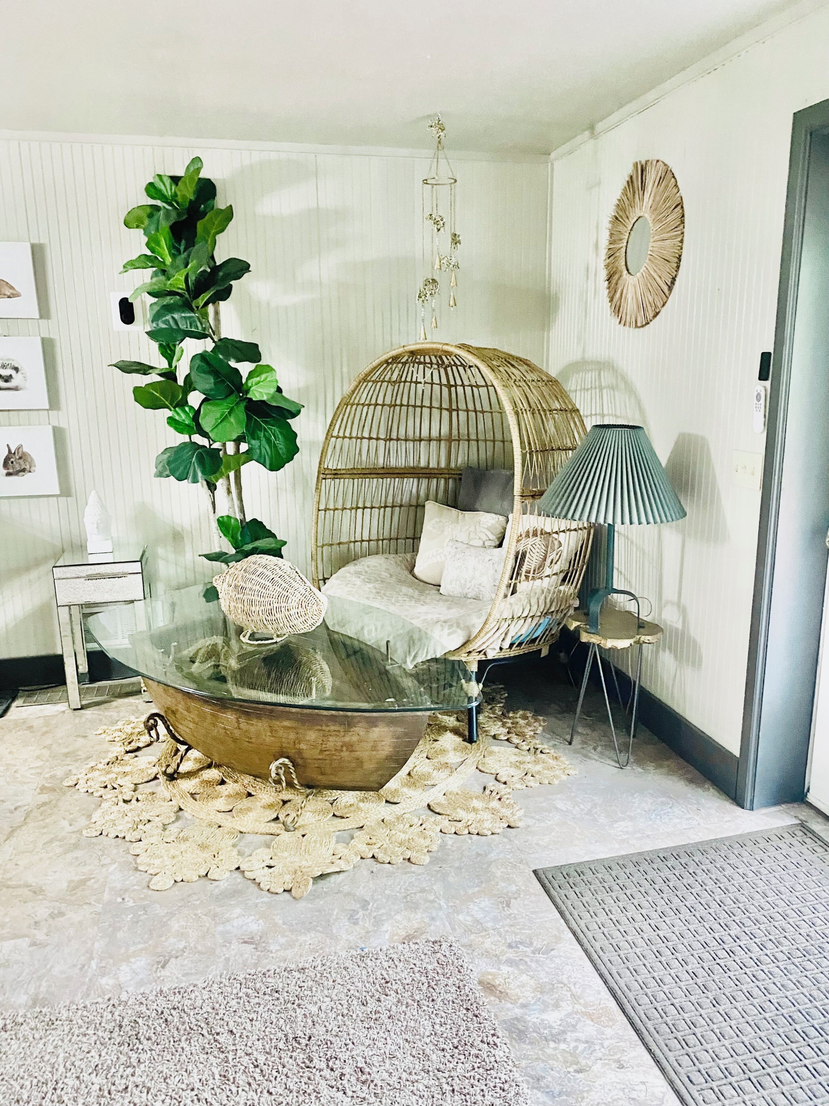 Un salon chaleureux avec une table basse en verre, un fauteuil en osier, une grande plante verte et un miroir décoratif.