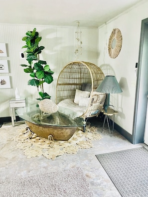 A cozy living room with a glass coffee table, a wicker chair, a large green plant, and a decorative mirror.